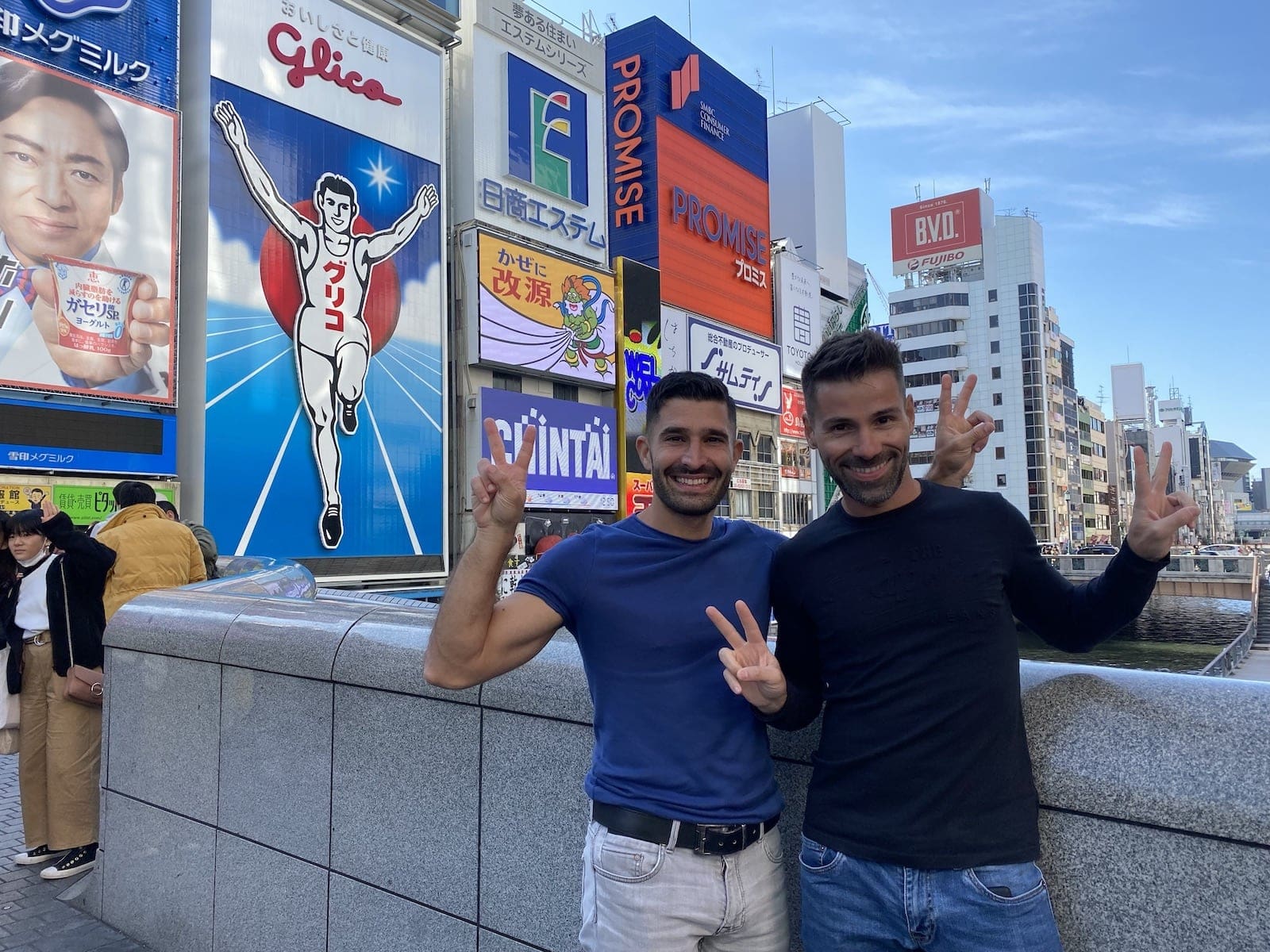 Gay travel bloggers, Nomadic Boys, give the peace sign in front of some very Japanese ads in Tokyo.